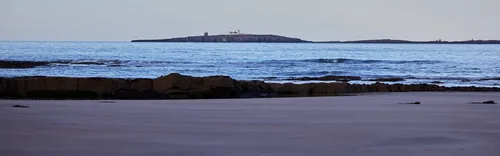 This landscape photograph shows the Farne Islands as viewed from a beach on the coast of Northumberland, England, United Kingdom. The image captures the scene on an autumn afternoon, with the water gently lapping against the rocky shoreline in the foreground. The Farne Islands are prominent in the distance, and lighthouses can be seen atop the islands, serving as recognizable landmarks. The clear sky and cool tones further emphasize the autumn season, highlighting the natural beauty of the beach and surrounding water.